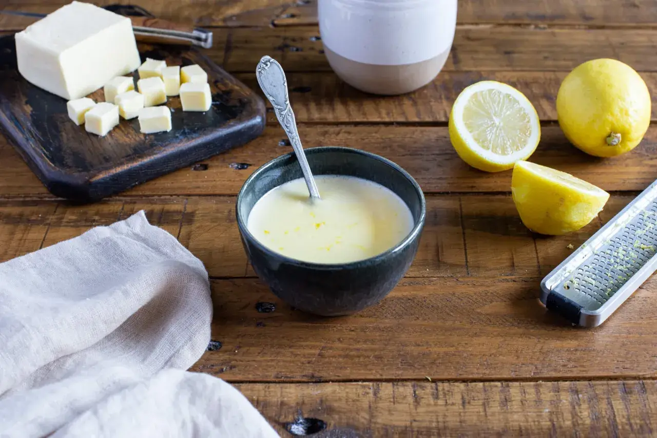 Un bol de sauce beurre citron, avec des citrons, du beurre coupé en dés et une râpe à zester sur une table en bois.