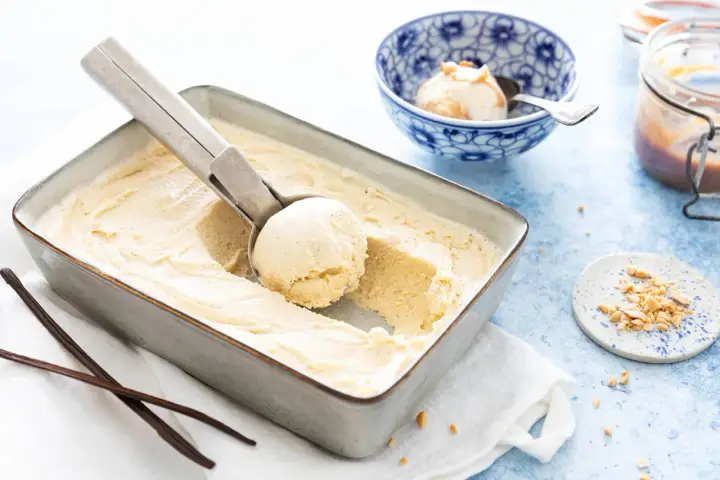 Une cuillère prélève une boule d'une glace vanille maison crémeuse dans un plat. Des gousses de vanille et des éclats de cacahuètes sont à proximité.