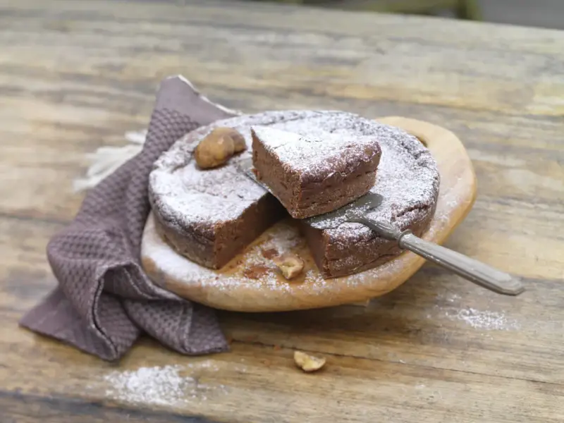 Gâteau au chocolat saupoudré de sucre glace, avec des châtaignes. Une part est coupée, prête à être dégustée. Une recette de châtaigne à essayer !